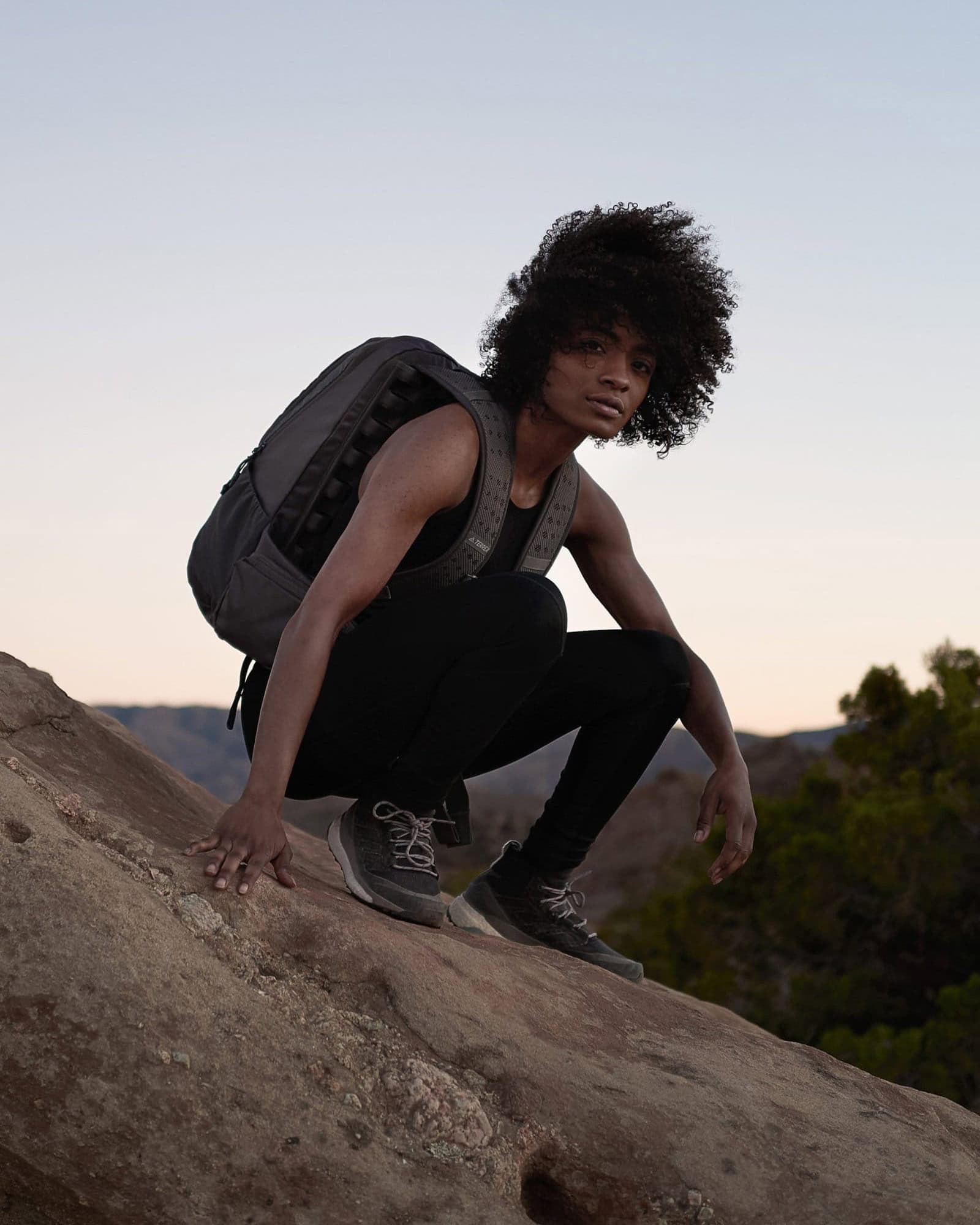 Young woman with curly hair, wearing a backpack, crouching on a rock during outdoor adventure at sunset.
