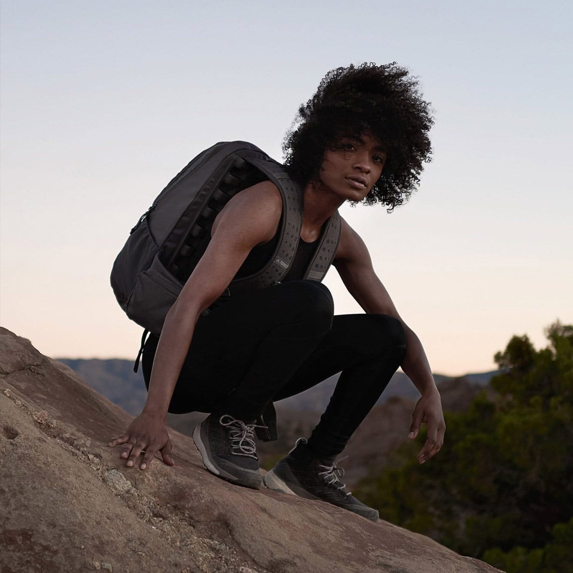 A young woman with natural curly hair crouching on a rocky terrain during sunset, wearing a backpack and outdoor clothing, ready for adventure.